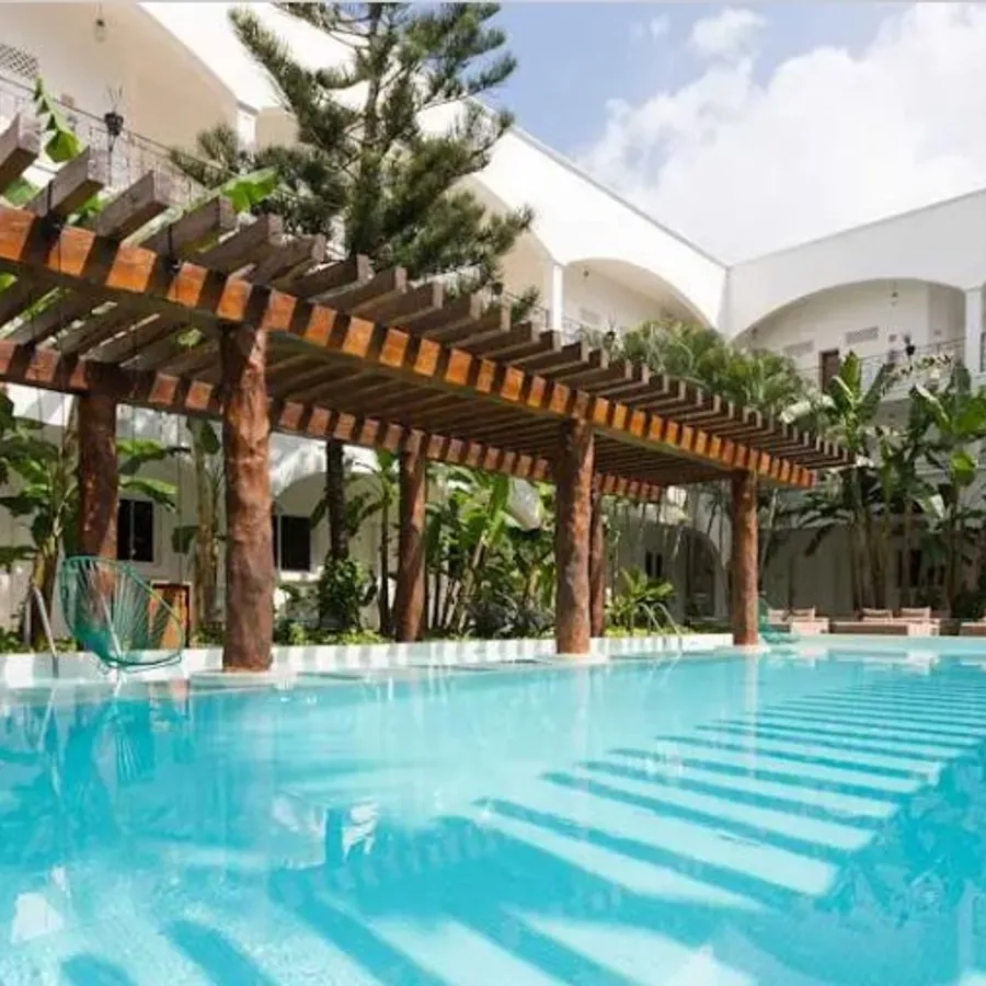 La photo montre une magnifique piscine extérieure entourée de chaises longues, située dans un cadre tropical avec des plantes exotiques et des palmiers. Un grand pergola en bois traverse la piscine, offrant une touche de charme rustique. En arrière-plan, le bâtiment de l