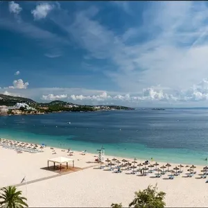 La photo montre une magnifique plage de sable fin bordée par des eaux turquoise, idéale pour un mariage à destination. Des parasols et des chaises longues sont soigneusement disposés, offrant un cadre idyllique pour les mariages organisés par Voyage Club Evasion, spécialiste du mariage à destination. À l'arrière-plan, un ciel bleu parsemé de quelques nuages et des bâtiments modernes complètent ce paysage paradisiaque, parfait pour célébrer un mariage élégant au Canada.