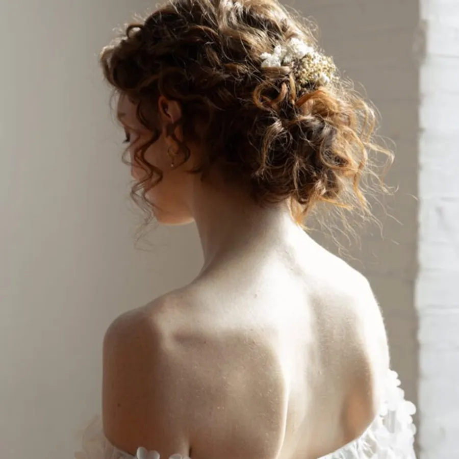 Une mariée de dos aux cheveux bouclés ornés de fleurs blanches, mise en valeur par Mariage MTL dans la catégorie Beauté pour le mariage. Elle porte une robe de mariée à épaules dénudées, créant une allure élégante et romantique. La lumière douce de la pièce illumine délicatement sa nuque.