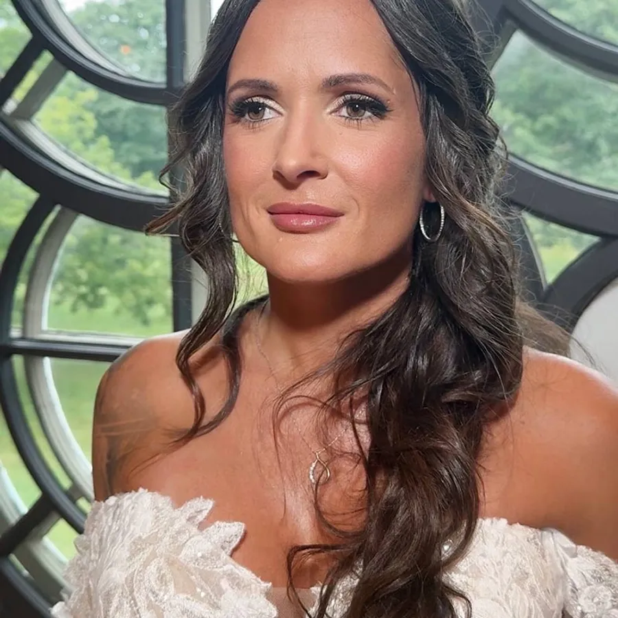 Femme portant une robe de mariée en dentelle blanche, avec des cheveux longs et ondulés et un maquillage subtil. Elle se trouve devant une grande fenêtre circulaire avec un paysage verdoyant visible en arrière-plan. Cette image illustre le travail de Mariage MTL dans la catégorie Beauté pour le mariage, soulignant le soin apporté à la coiffure et au maquillage pour sublimer la mariée le jour de son mariage.