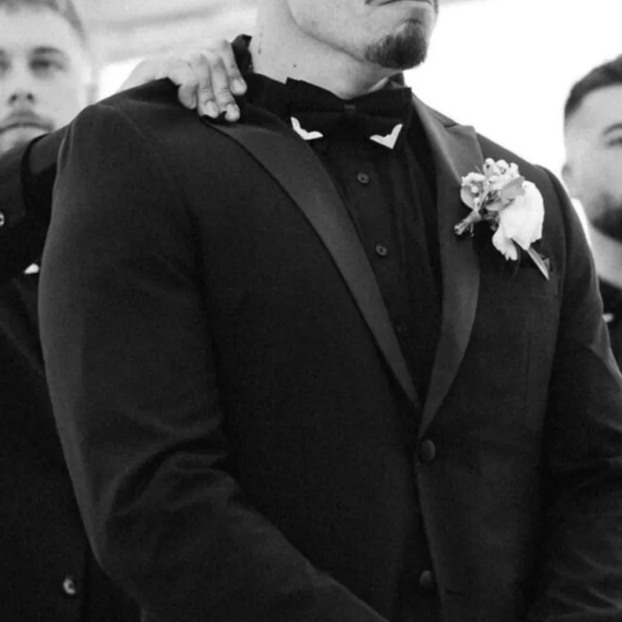 A groom elegantly dressed in a classic black tuxedo, provided by M/2 Boutiques, stands with a solemn expression at a wedding ceremony. The tuxedo features satin lapels, a matching black shirt, and a stylish bow tie, emphasizing a refined look ideal for formal weddings. A boutonnière of white flowers adorns the lapel, adding a touch of sophistication. Another person is partially visible beside him, resting their hand supportively on his shoulder. This image highlights the Wedding Tuxedo offering by M/2 Boutiques, fitting perfectly within the Mariage Elegant vendor listings in Canada.