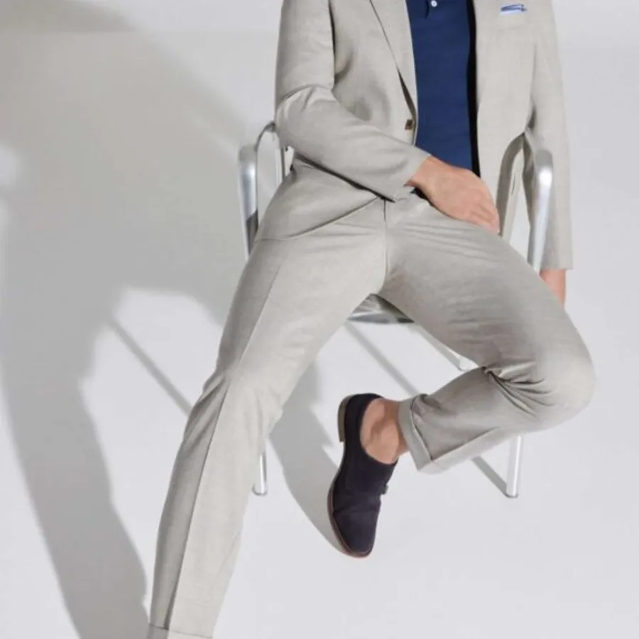 A stylish model is seated on a modern metal chair, wearing an elegant, light grey tuxedo by M/2 Boutiques, perfect for weddings. The tuxedo includes a tailored jacket and matching trousers, paired with a dark blue dress shirt. The ensemble is completed with dark shoes, adding a touch of contrast. The setting is minimalistic, emphasizing the sophisticated design of the tuxedo, ideal for grooms looking for a contemporary wedding outfit. This image represents the wedding tuxedo category on Mariage Elegant, Canada, showcasing vendors like M/2 Boutiques.