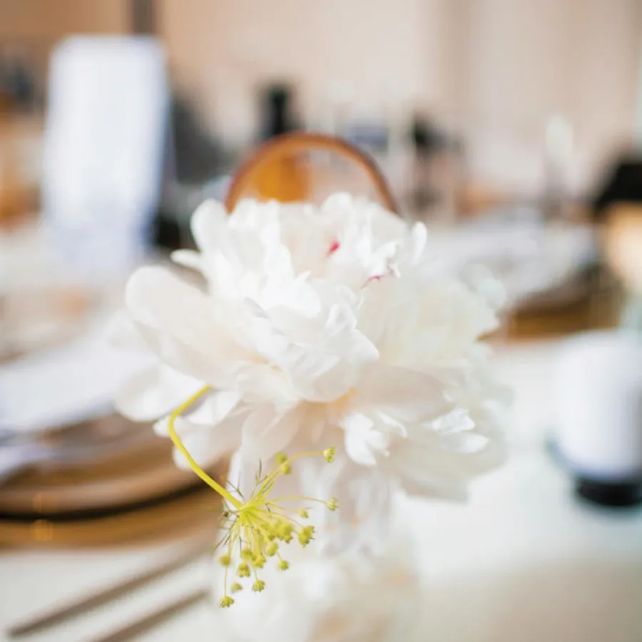A beautifully arranged centerpiece featuring large, soft white peonies and delicate greenery sits prominently on a elegantly set table, creating a serene and elegant ambiance. The background shows slightly blurred dining settings, including polished cutlery and glassware, suggesting a sophisticated wedding reception. This image, captured by Inhouse Studios Photography, exemplifies the artistry in the Wedding Photography category, making it a stunning representation for Mariage Elegant in Canada.